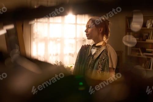 Preview: Young Woman Standing by Window Holding Clothes in Sunlit Room