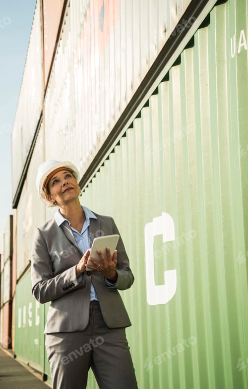 Preview: Businesswoman with digital tablet wearing safety helmet at container port