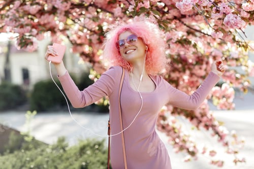 Preview: Happy woman with curly pink hair near the blossoming spring tree listening music on smartphone.