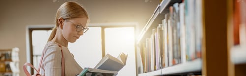 Preview: Smart female student in glasses reads book while standing in the university library