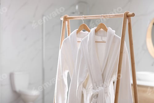 Preview: Fresh white bathrobes hanging on rack indoors