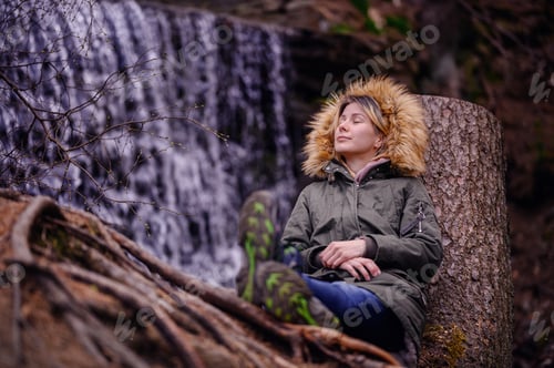 Preview: Young woman sits leaning on a stump in green parka with fur before waterfall alone in forest