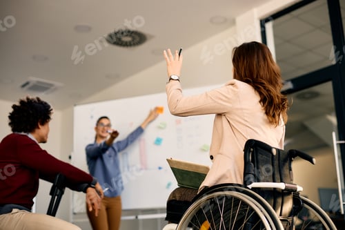Preview: Rear view of businesswoman in wheelchair raising arm to ask question during presentation in office.