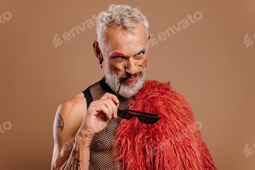 Preview: Stylish mature gay man with colored eyebrows holding glasses against brown background