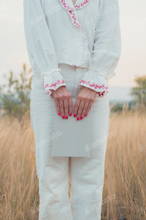 Preview: Standing woman dressed in white holds a book on a meadow at sunset