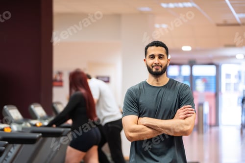 Preview: Relaxed man with arms crossed in gym, people behind.