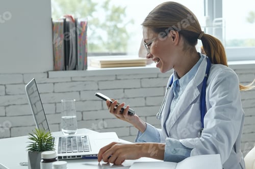 Preview: Confident female doctor using smart phone and smiling while sitting at the medical office