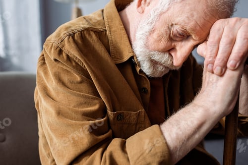 Preview: senior, depressed man sitting with bowed head while leaning on walking stick