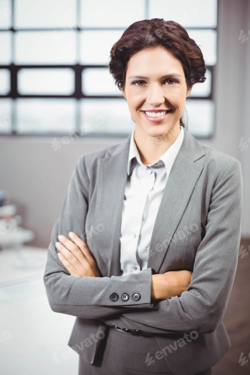 Preview: Businesswoman wearing grey suit posing crossing arms at desk with monitor and grid window in office
