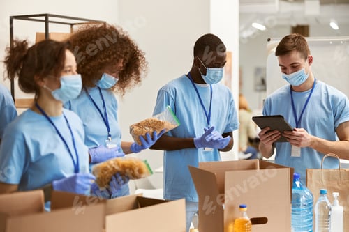 Preview: Team of volunteers in protective masks and gloves sorting, packing food in cardboard boxes, working