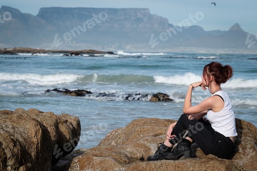 Preview: Young woman in a white top and black pants sitting on a boulder next to the ocean