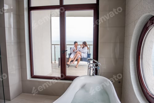 Preview: Couple in white bathrobes are sitting on open terrace by the sea, drinking tea with lemon from cups