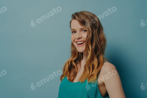Preview: Positive millenial girl with wavy hair stands sideways and looks at camera with joyful radiant smile