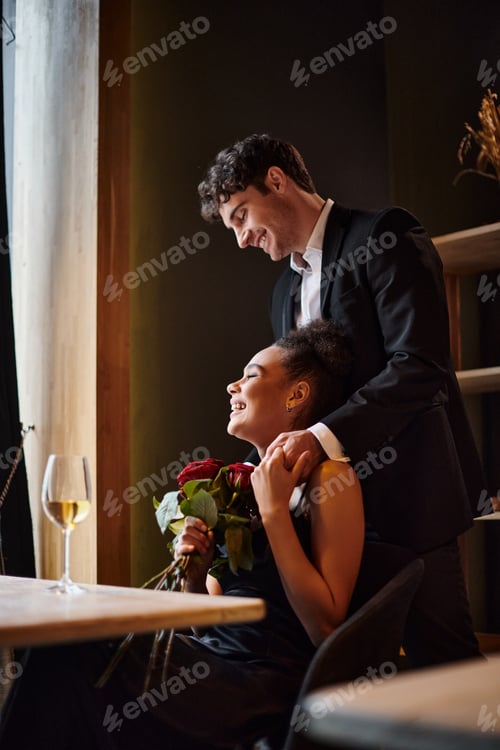 Preview: happy african american woman holding hands with man in suit standing behind her, red roses