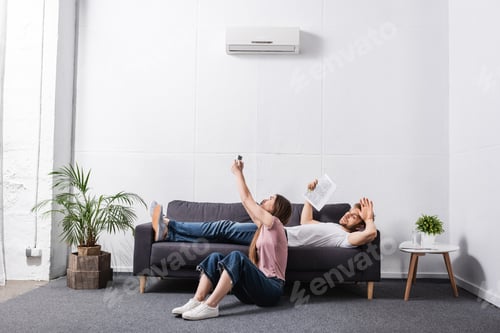Preview: young couple with remote controller and newspaper suffering from heat while sitting home with broken