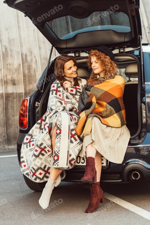 Preview: happy adult women wrapped in blankets sitting in car trunk at urban street