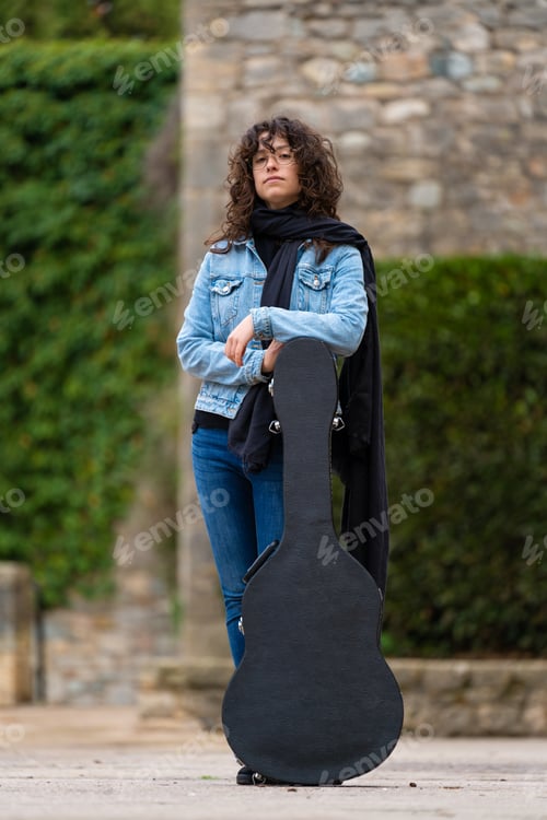 Preview: Young woman with curly hair and glasses posing with a guitar case in the street