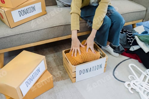Preview: Asian young woman packing clothes at home, putting on stuff into donate box with second hand clothes