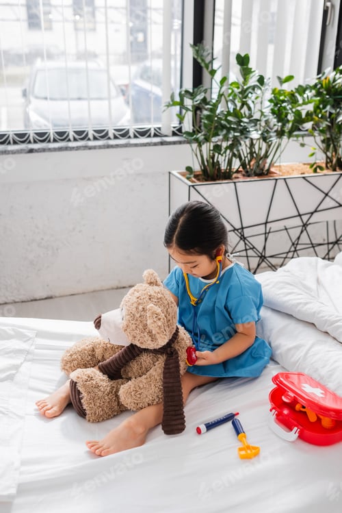 Preview: asian child in hospital gown examining teddy bear with toy stethoscope on bed in clinic
