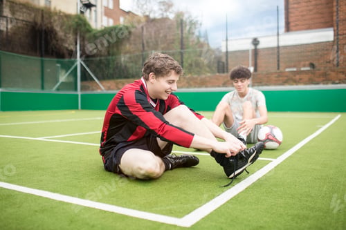 Preview: Two young men on urban football pitch, tying shoelaces