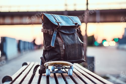 Preview: Close-up photo of a backpack lying on a skateboard that stands on a bench