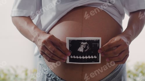 Preview: Young Asian Pregnant women show and looking ultrasound photo baby in belly.