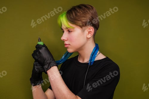 Preview: Young man tattoo artist with colored hair and piercing holding machine indoor. Small business