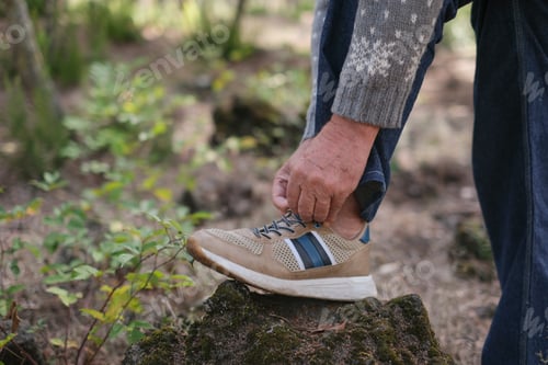 Preview: Senior man bends over to tie his shoes outdoors, enjoying nature in a forest park on a sunny day