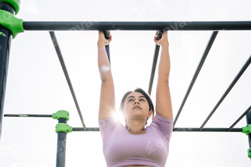 Preview: Motivated young ethnic sportswoman exercising on high bars in sunlight