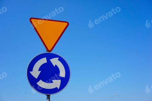 Visualização: Foto aproximada de uma placa de trânsito avisando sobre uma próxima rotatória sob um céu azul claro