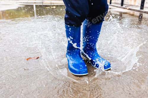 Preview: Young boy with blue rubber boots jumps over a puddle of water in a park in his city.