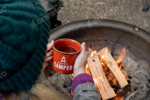 Preview: Camping lifestyle concept. Girl holding red enamel mug with tea near campfire. Happy camper