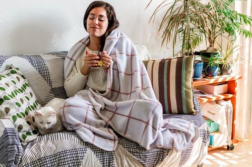 Preview: Young female wrapped in a blanket sitting on a sofa with a white cat and drinking a cup of tea