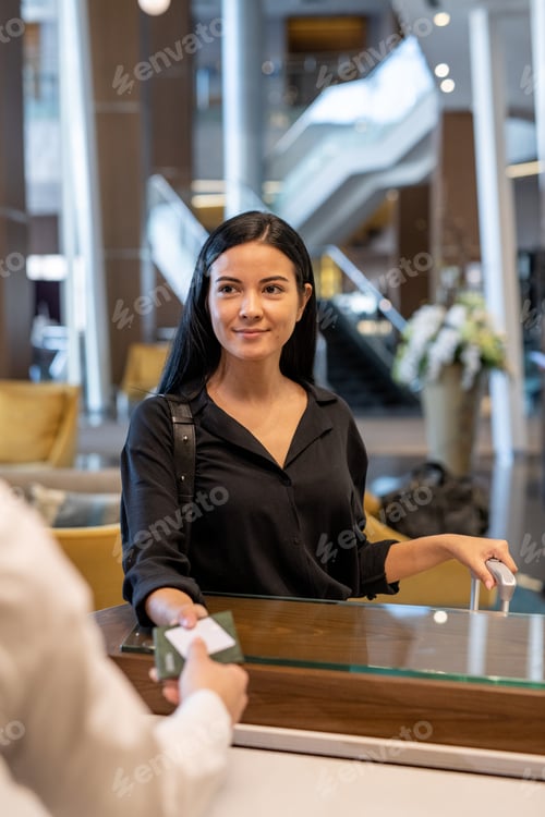 Preview: Happy young businesswoman or female traveler with luggage standing by reception counter