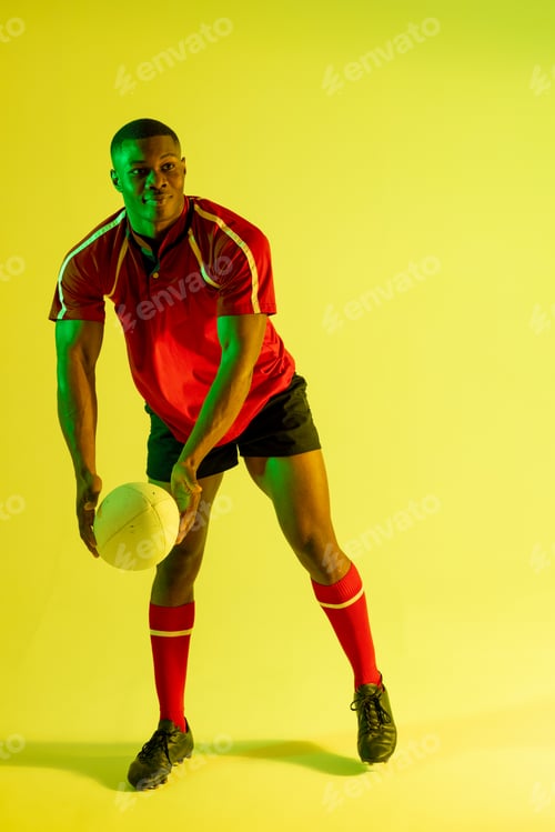 Preview: African american male rugby player with rugby ball over yellow lighting