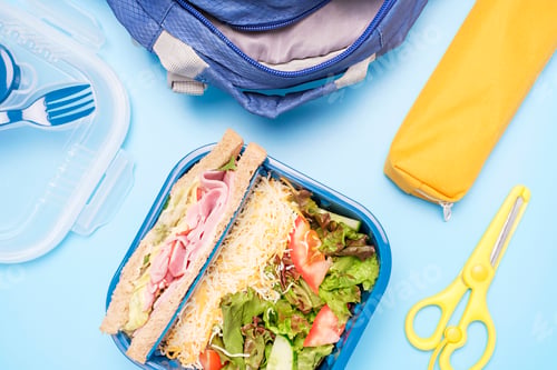 Preview: Backpack and plastic lunchbox with sandwich and salad on blue background top view.
