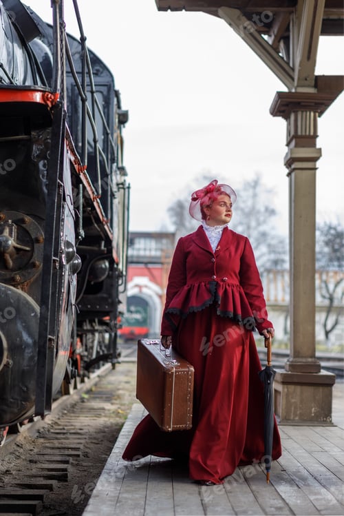 Preview: A girl in a burgundy suit and a hat stands with a suitcase near an old steam locomotive.Vintage