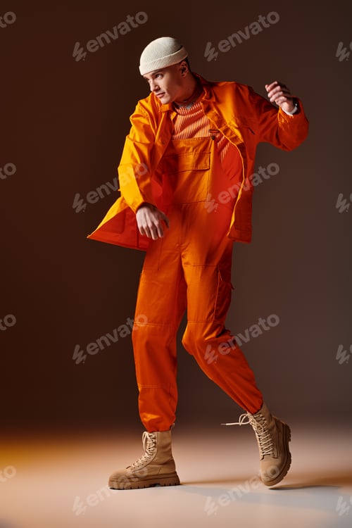Preview: Full length shot of man in orange jumpsuit and jacket, beige beanie posing on brown background