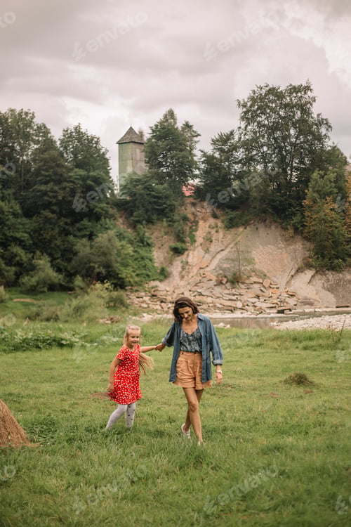 Preview: A little girl in a red dress walks with her mother on the green grass near the river