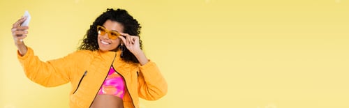 Preview: happy african american young woman adjusting sunglasses while taking selfie isolated on yellow,