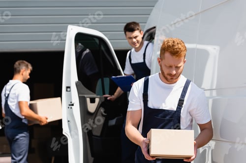 Preview: Selective focus of loader holding cardboard box while colleagues working near truck on urban street