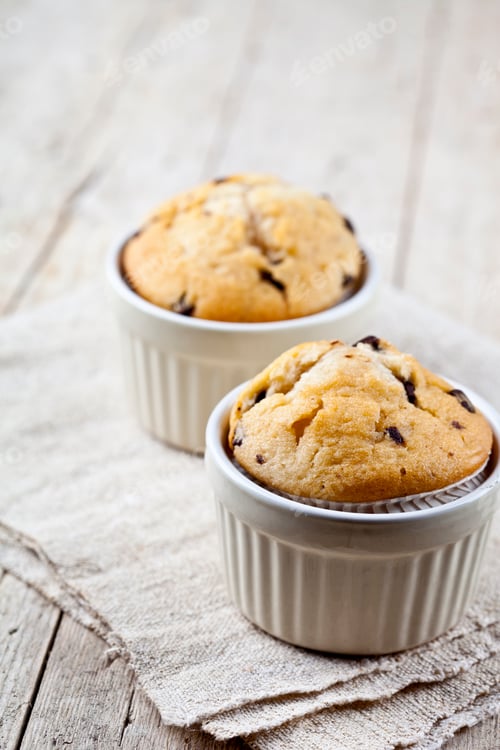 Preview: Two homemade fresh muffins on ceramic white bowls on linen napkin on rustic wooden table.