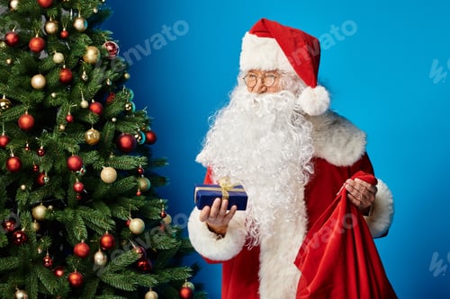 Preview: happy Santa Claus with beard and eyeglasses in red costume holding sack bag and Christmas present