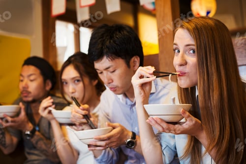 Preview: Four people sitting sidy by side at a table in a restaurant, eating from bowls using chopsticks.