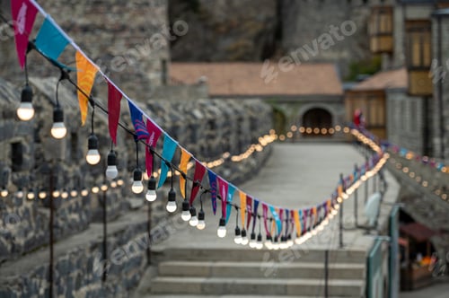 Preview: Garland of colorful flags and glowing light bulbs against a defocused background of castle.