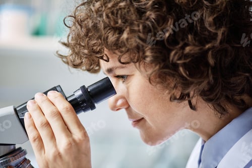Preview: Side view of young serious brunette female biochemist looking in microscope