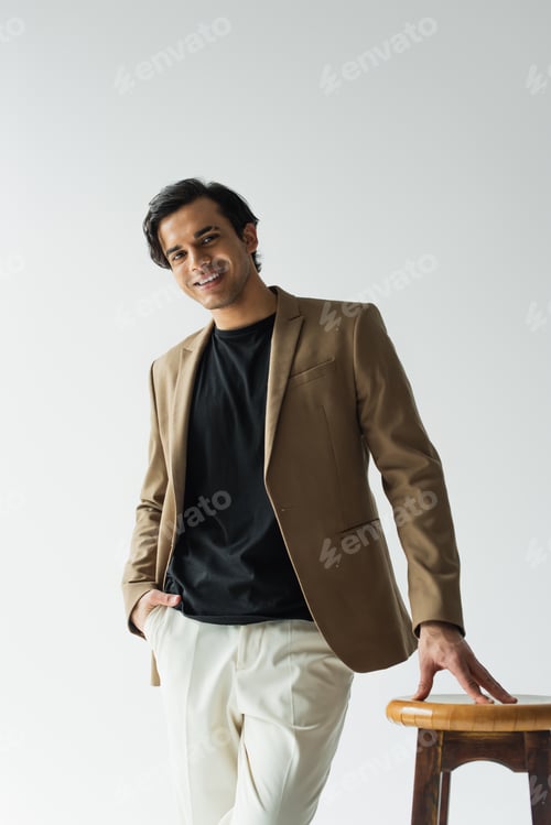 Preview: happy young man in stylish beige blazer posing with hand in pocket near wooden chair isolated on