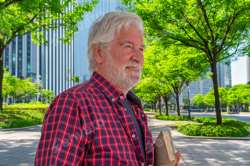 Preview: Handsome senior bearded man walking in urban street with a book under his arm.