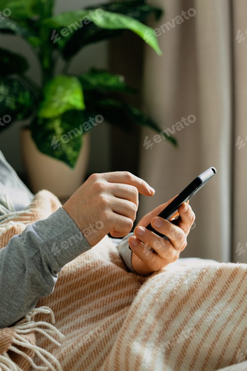 Preview: a caucasian man relaxing, using smart phone lying in bed under throw at home