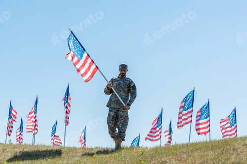 Preview: handsome soldier in military uniform and cap holding american flag while standing on grass